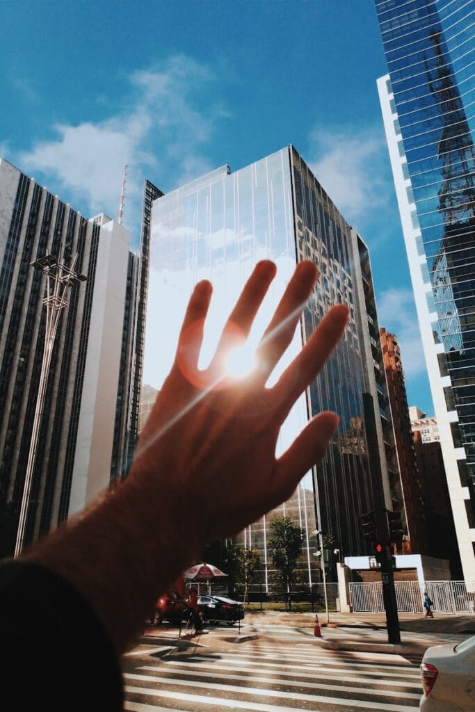 A hand reaches towards modern glass skyscrapers reflecting sunlight in a vibrant urban setting.